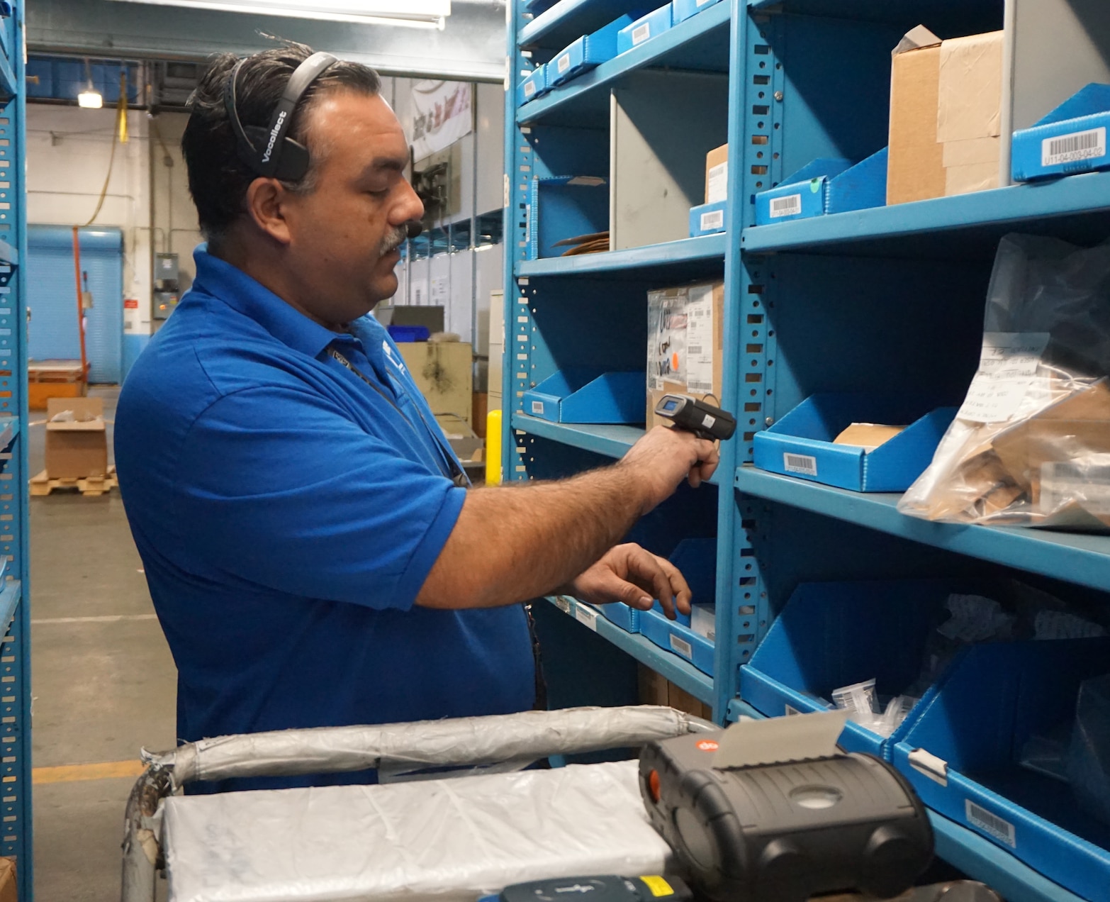 A man uses a headset and a small device attached to his hand to select items from a warehouse shelf
