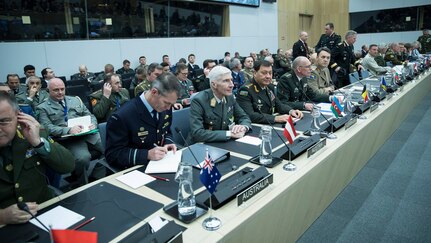 Military officers from various nations sit behind microphones at a round table with lothers seated behind them.