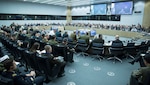 Wide-angle shot of a large conference facility, dominated by a round table with military officers seated around it.