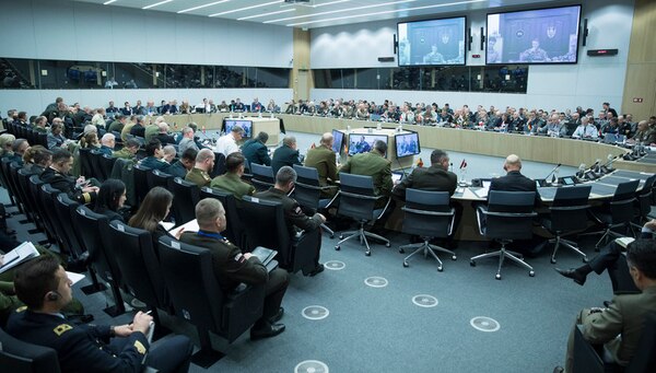 Wide-angle shot of a large conference facility, dominated by a round table with military officers seated around it.