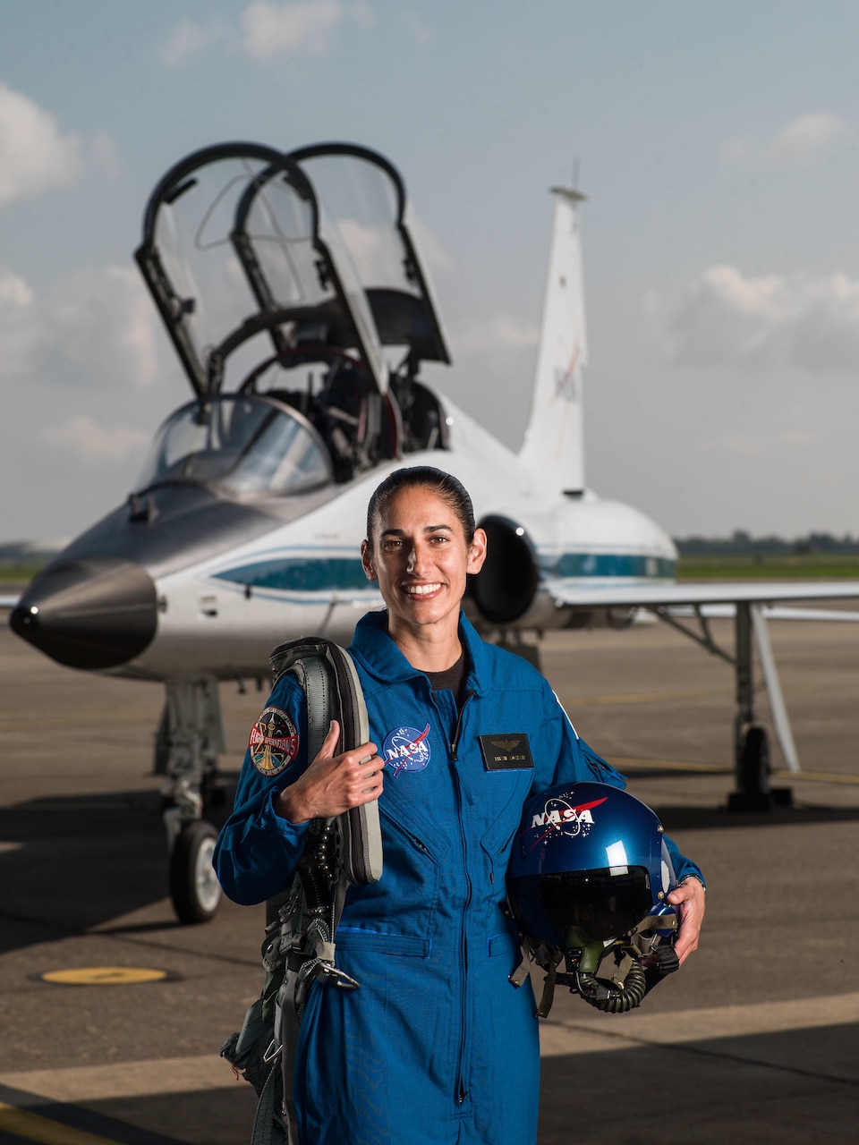 A female astronaut wearing a blue flight suit and NASA-logoed helmet poses in front of a T-38 Talon trainer airplane.