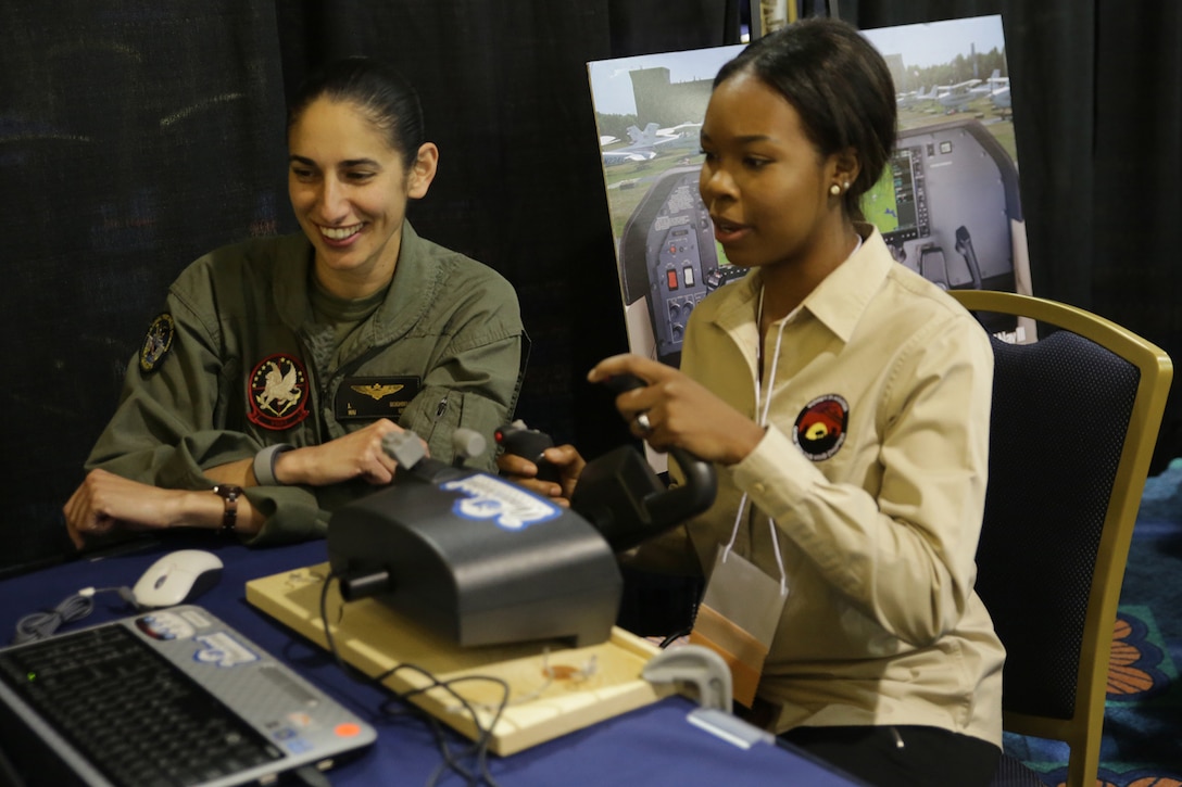 A pilot in flight suit sits beside a teenager who’s using the joystick of a computer simulator. Both are watching a screen.