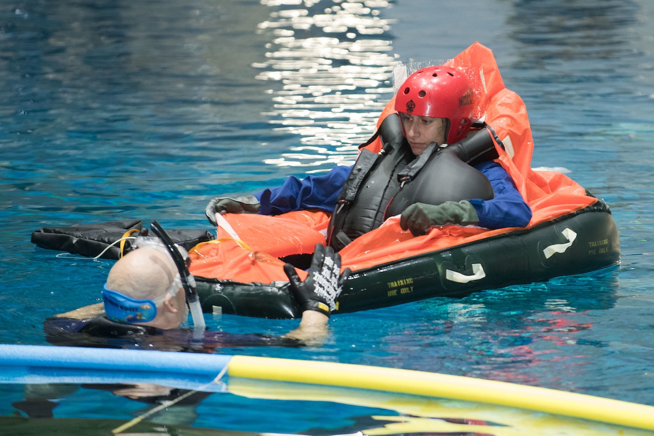 A woman wearing a large blow-up vest and helmet sits in an inflatable in a pool while taking instruction from a man in the water in a diving suit with snorkel.