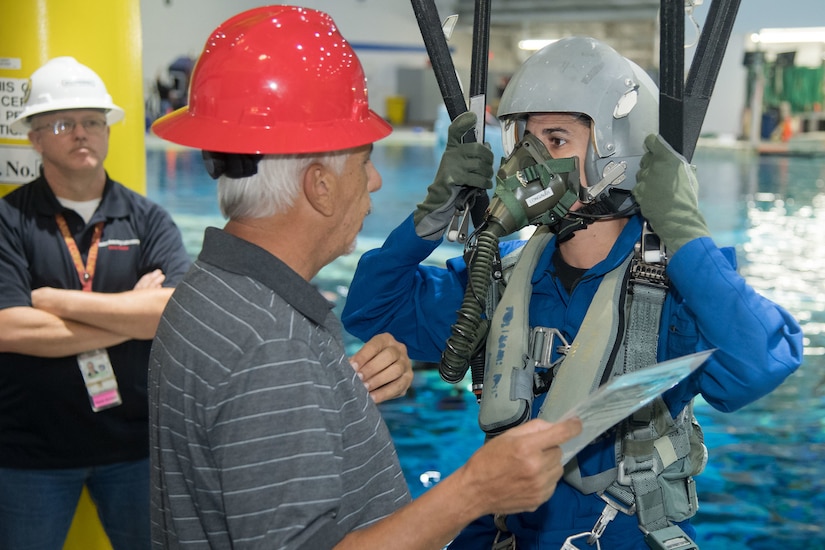 A trainee wearing a mask, helmet and what appears to be parachute cables listens to a man wearing a red hardhat. A pool is in the background.