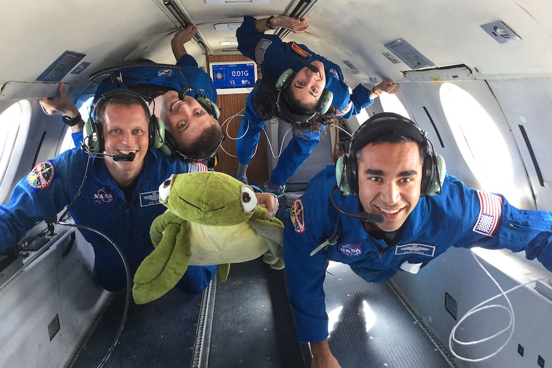 Four astronaut candidates in flight suits smile at the camera as they float in a reduced gravity environment on an airplane.