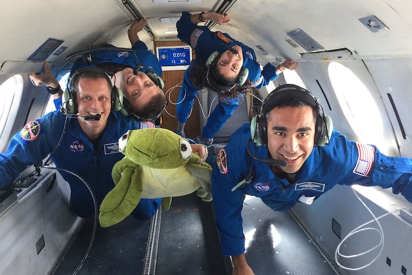 Four astronaut candidates in flight suits smile at the camera as they float in a reduced gravity environment on an airplane.