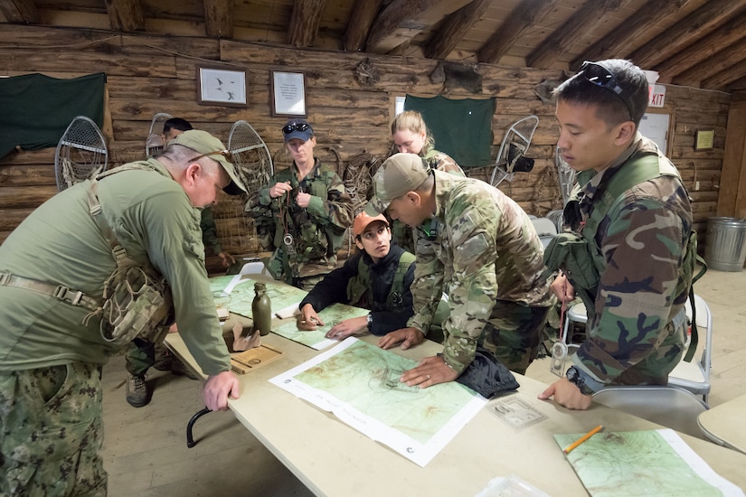 Seven people in military camouflage sit and stand around a table looking at maps in what appears to be a wooden log cabin.