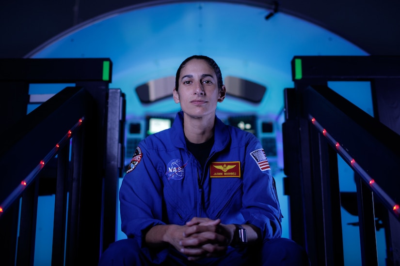 An astronaut in a blue flight suit sits on darkened steps, clasping her hands. A blue light illuminates a control room in the background.