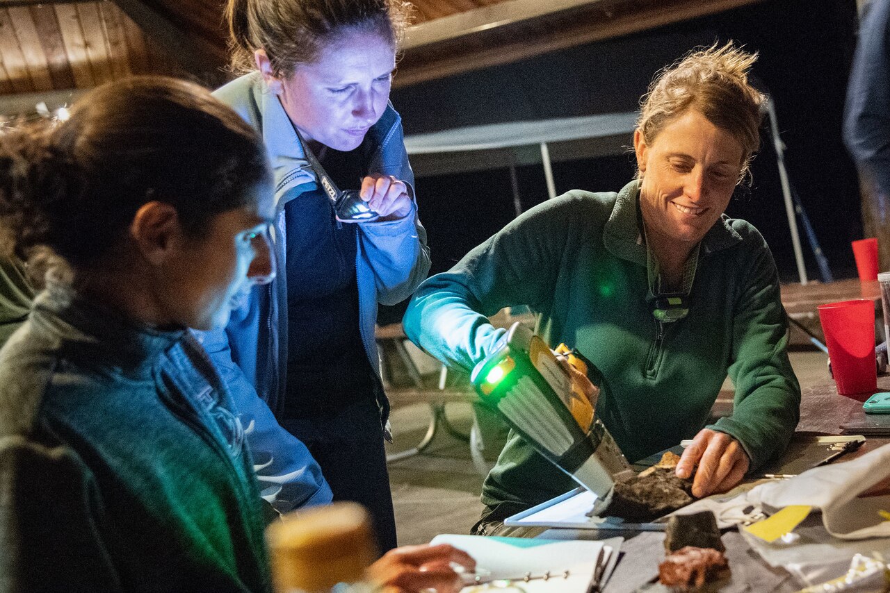 Three women huddle around a rock as they sit at an outdoor picnic table at night. One holds a flashlight while a second scans a rock on the table with a device.