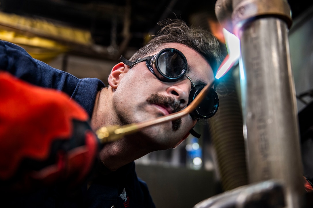 A sailor wearing goggles  uses a blowtorch to heat a pipe.