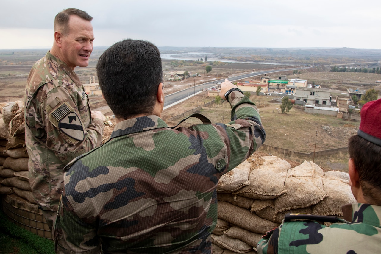 Two men in camouflage uniforms stand near a sandbag wall looking out over a town.