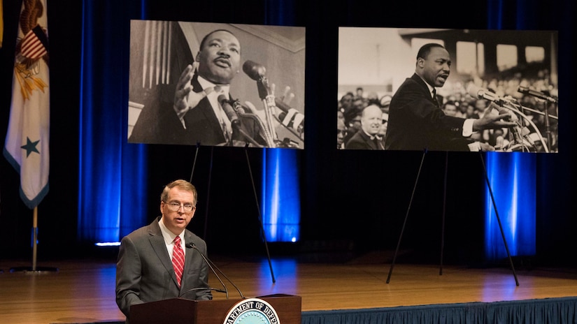 A man speaks from behind a lectern with large pictures behind him.