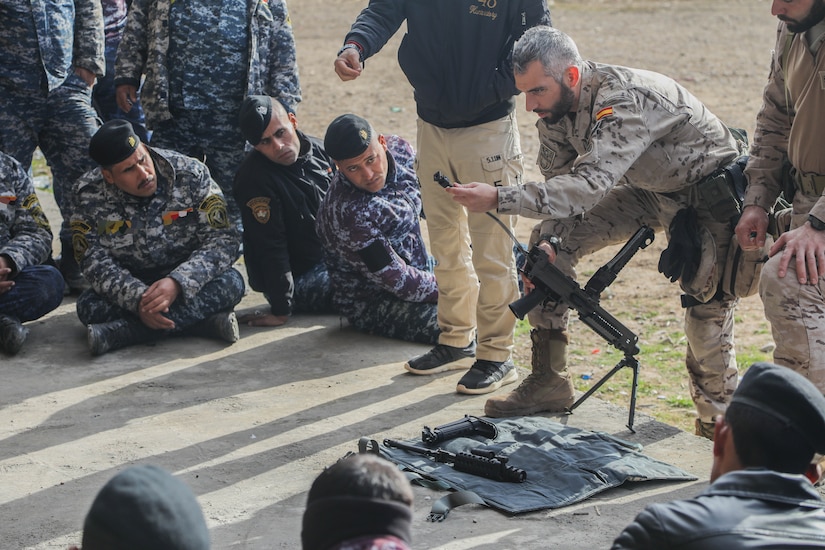 A man in a camouflage uniform holds a rifle, in pieces, while other men observe.