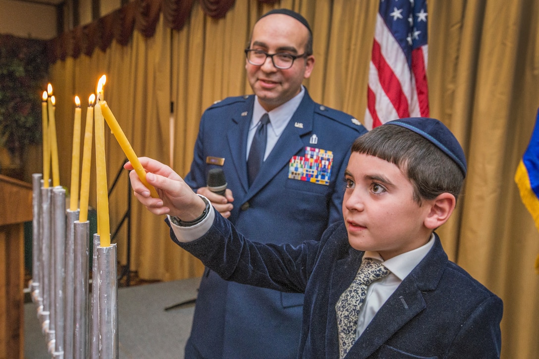 A chaplain observes as a child lights a candle on a menorah.