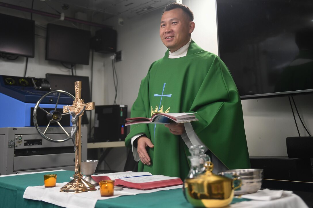 A man in religious garb holds a book.  On the table in front of him is a crucifix.