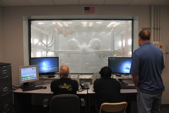 Three technicians around a desk monitor computers and look through a window in front of them where and A-10 aircraft is being depainted by two robots flanking the aircraft.