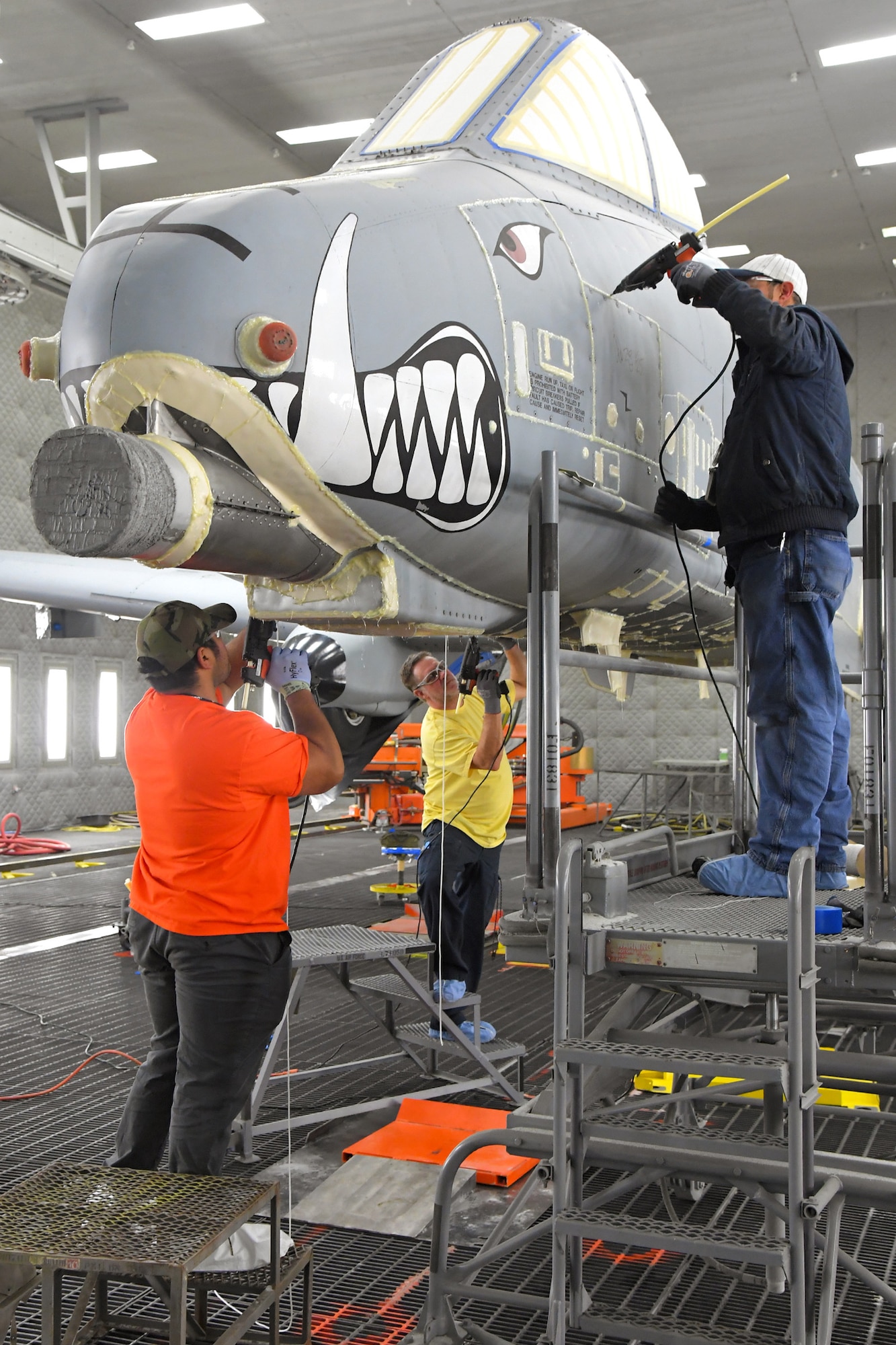 Three technicians work on the nose of an A-10 aircraft in a lighted depaint facility.