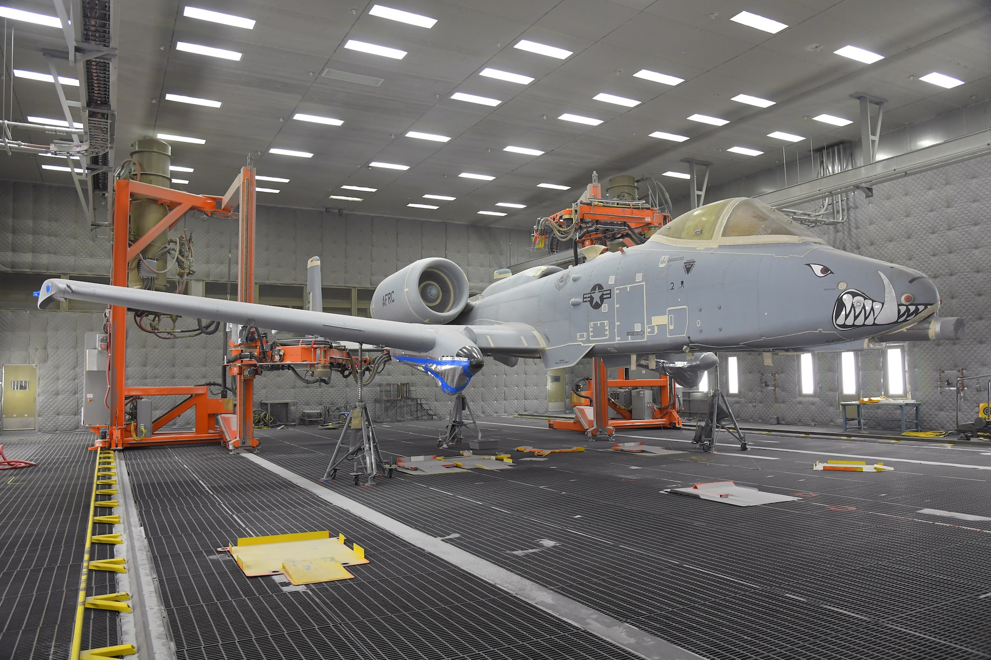 An A-10 aircraft sits on jacks in a lighted depaint facility with two orange depainting robots flanking it in the background.