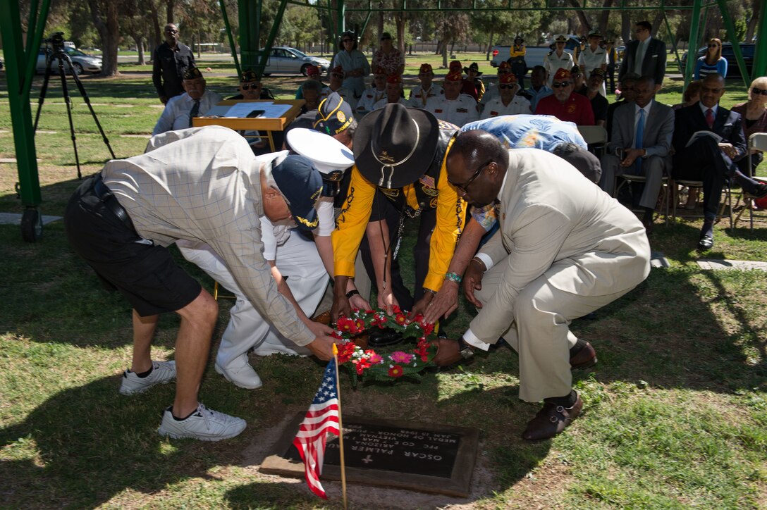Five older gentlemen lay a wreath at a gravestone as several veterans seated in the background look on.