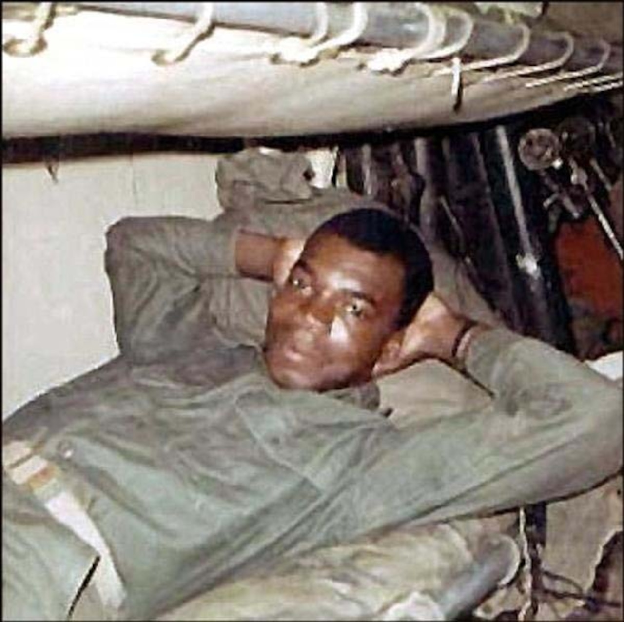 A young Marine in utility uniform relaxes in a bunk with his hands behind his head.