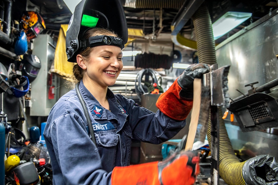 A sailor with orange gloves holds a silver belt for welding.