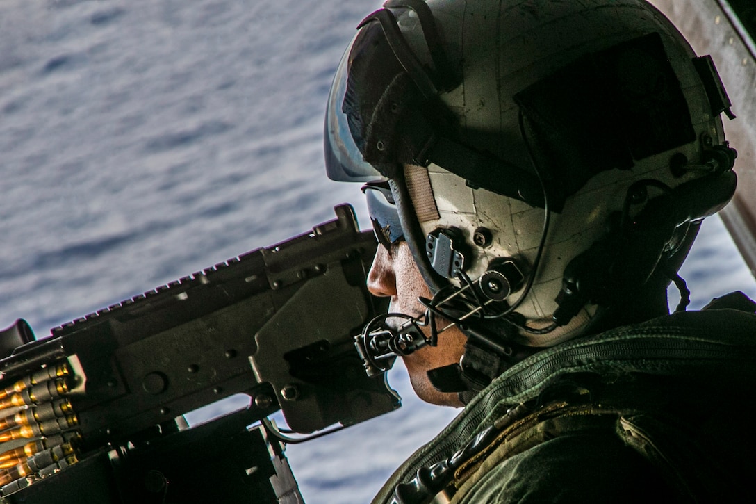 A Marine wears a helmet and sunglasses while in an aircraft over the sea.