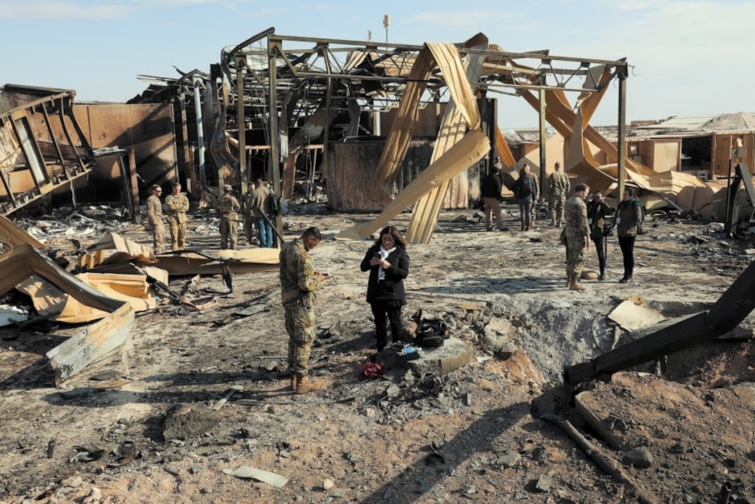 Soldiers and civilians stand on the site of a destroyed metal pole-barn-style building.
