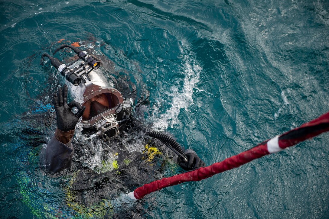 A sailor wearing dive gear while submerged in water gives a hand signal.