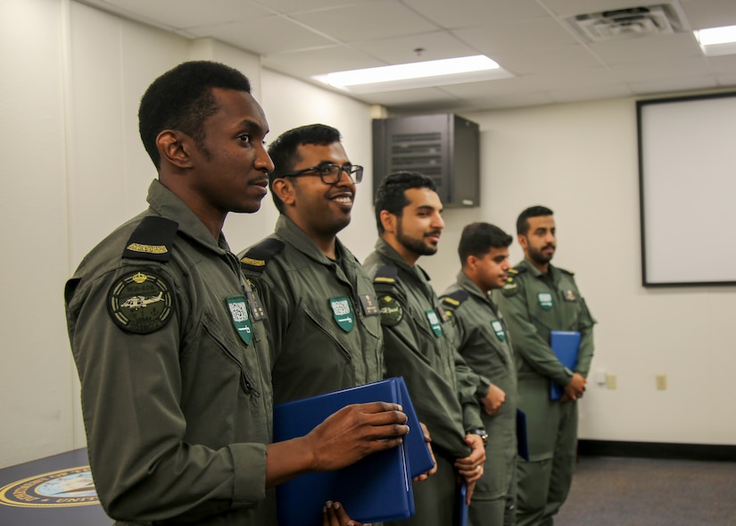 Five military personnel stand in line, each holds a blue folder.