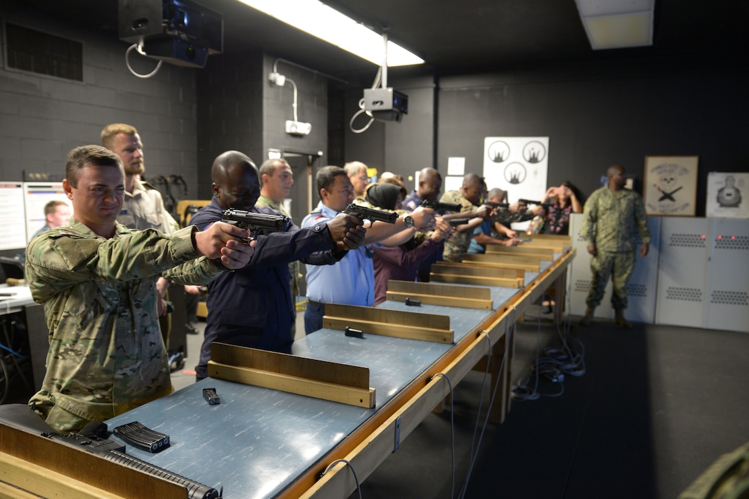 A line of military personnel stand behind a long table and fire hand guns.