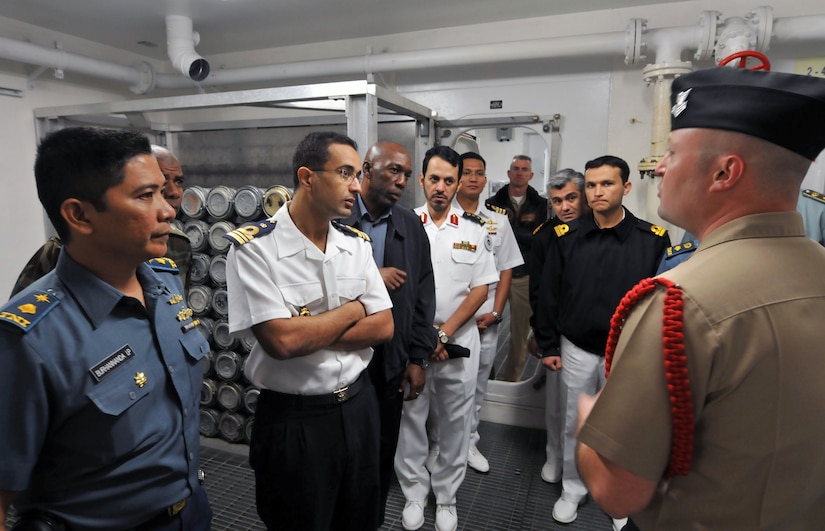 A group of military personnel, dressed in a variety of uniforms, stand aside each other while listening to another service member speak.