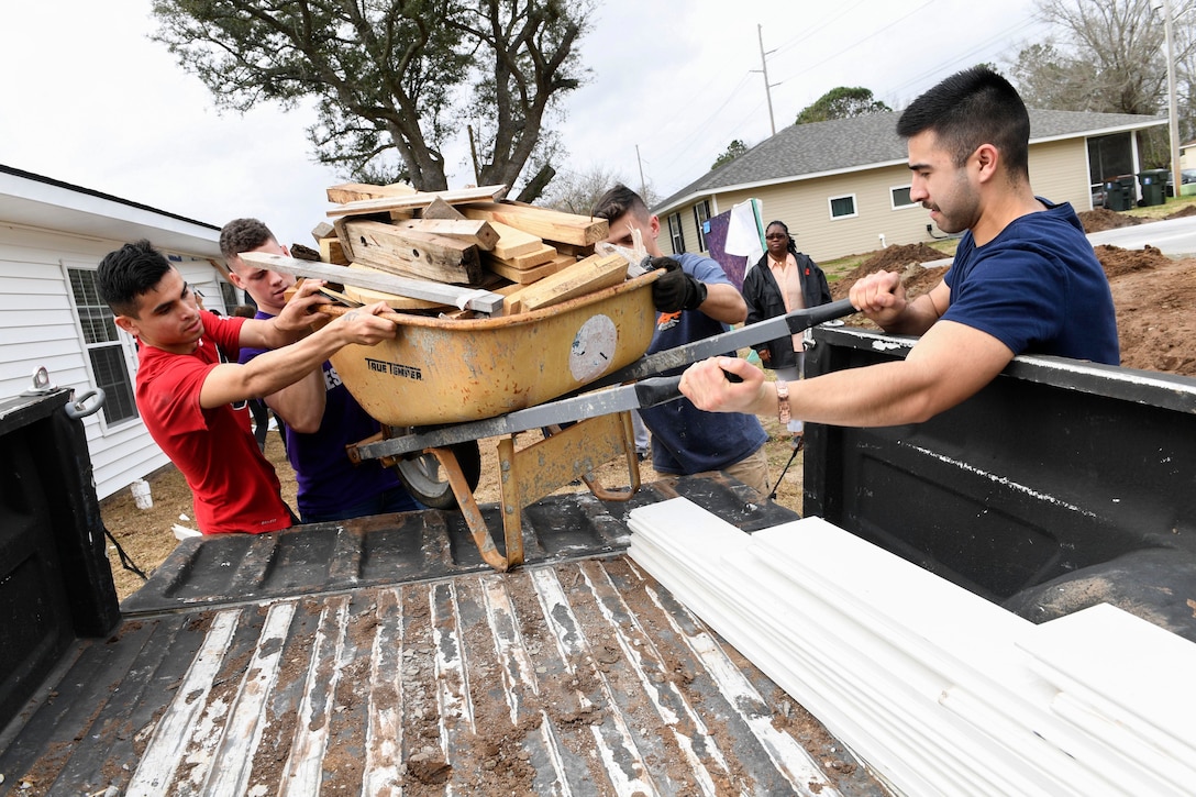 A group of sailors lift a wheelbarrow of wood from the back of a truck.