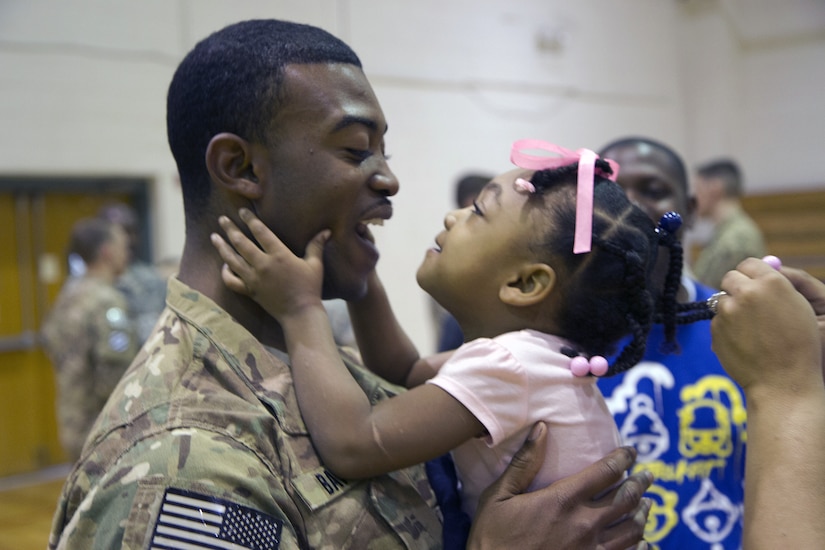 A little girl wearing a pink shirt and hair bow holds her smiling dad's face as they embrace.