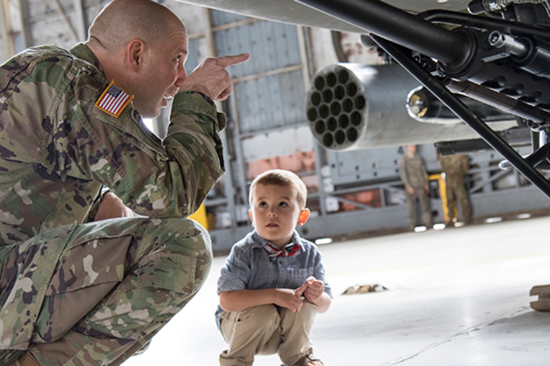 A squatting soldier points to something on the bottom of an airplane as a little boy, also squatting, looks at the soldier.