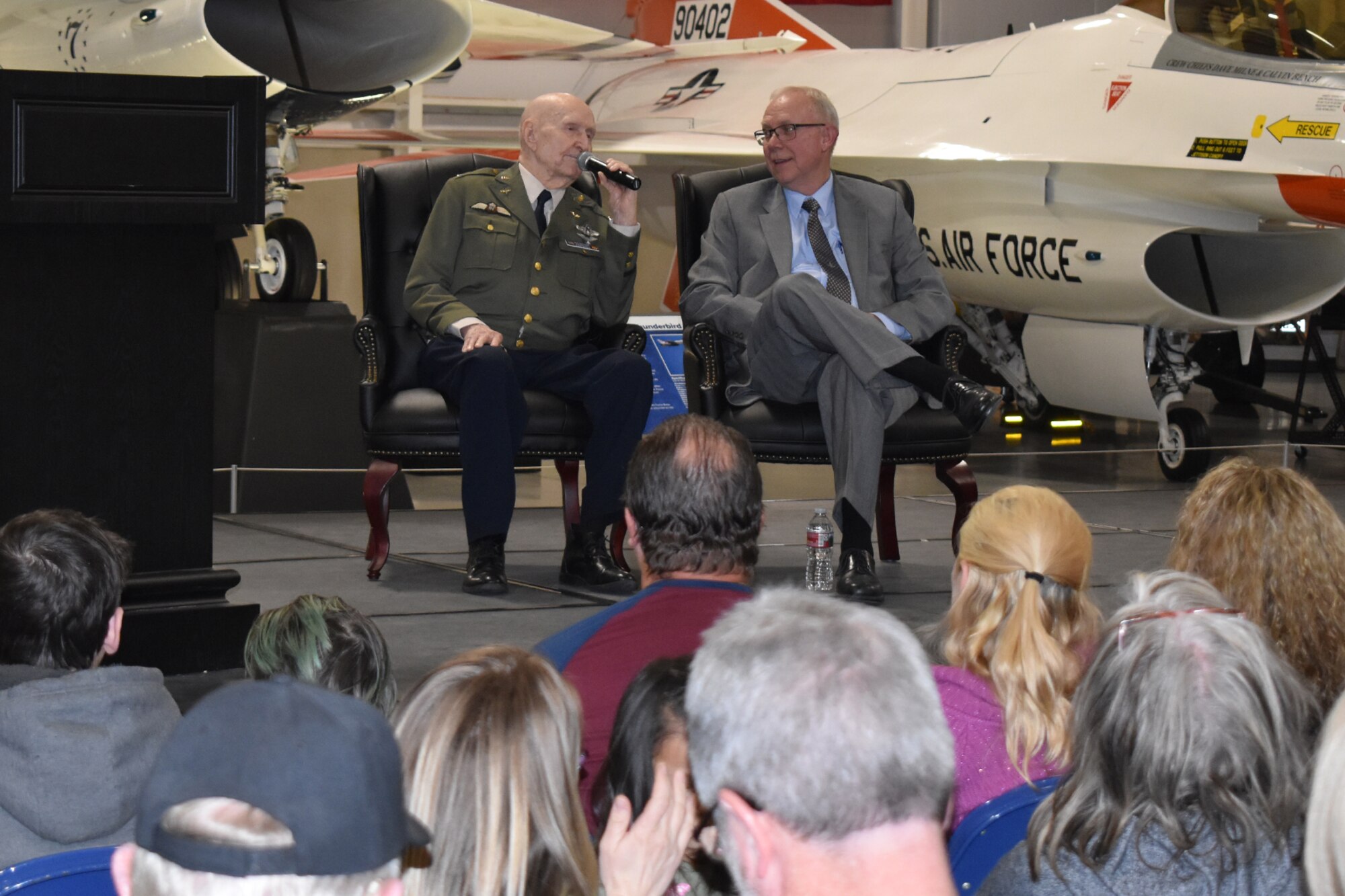 (Left to right) Retired Air Force Col. Gail Halovrsen  next to James Stewart, executive director of the Gail S. Halvorsen Aviation and Education Foundation, in chairs on a stage positioned in front of a static display of a U.S. Air Force Thunderbirds-painted F-16 inside the Hill Aerospace. Part of an audience watches on in the foreground.