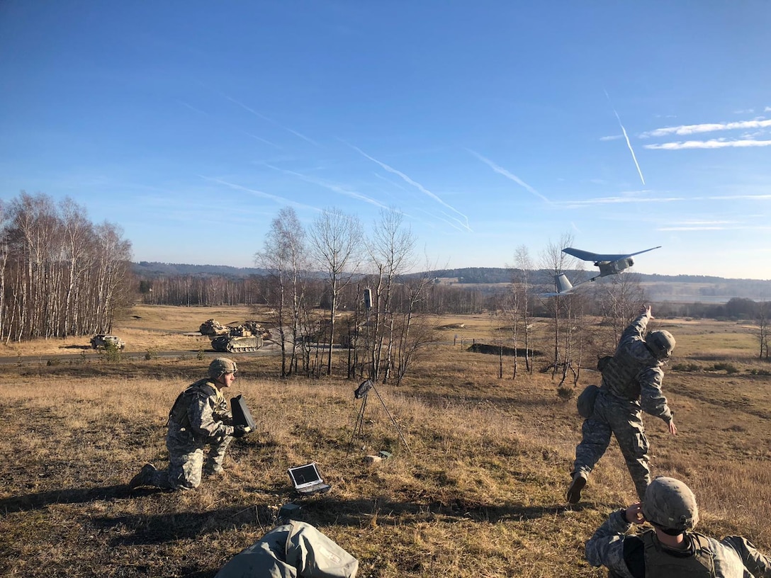 A soldier kneels on the ground as another throws an aerial device.