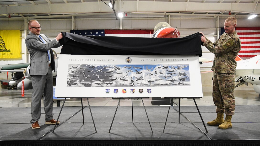 Aaron Clark at left and Col Jon Eberlan at right lift a black cloth from the top of a over-sized version of the 80th anniversary commemorative lithograph depicting the major weapons systems that have resided at Hill Air Force Base over the years.