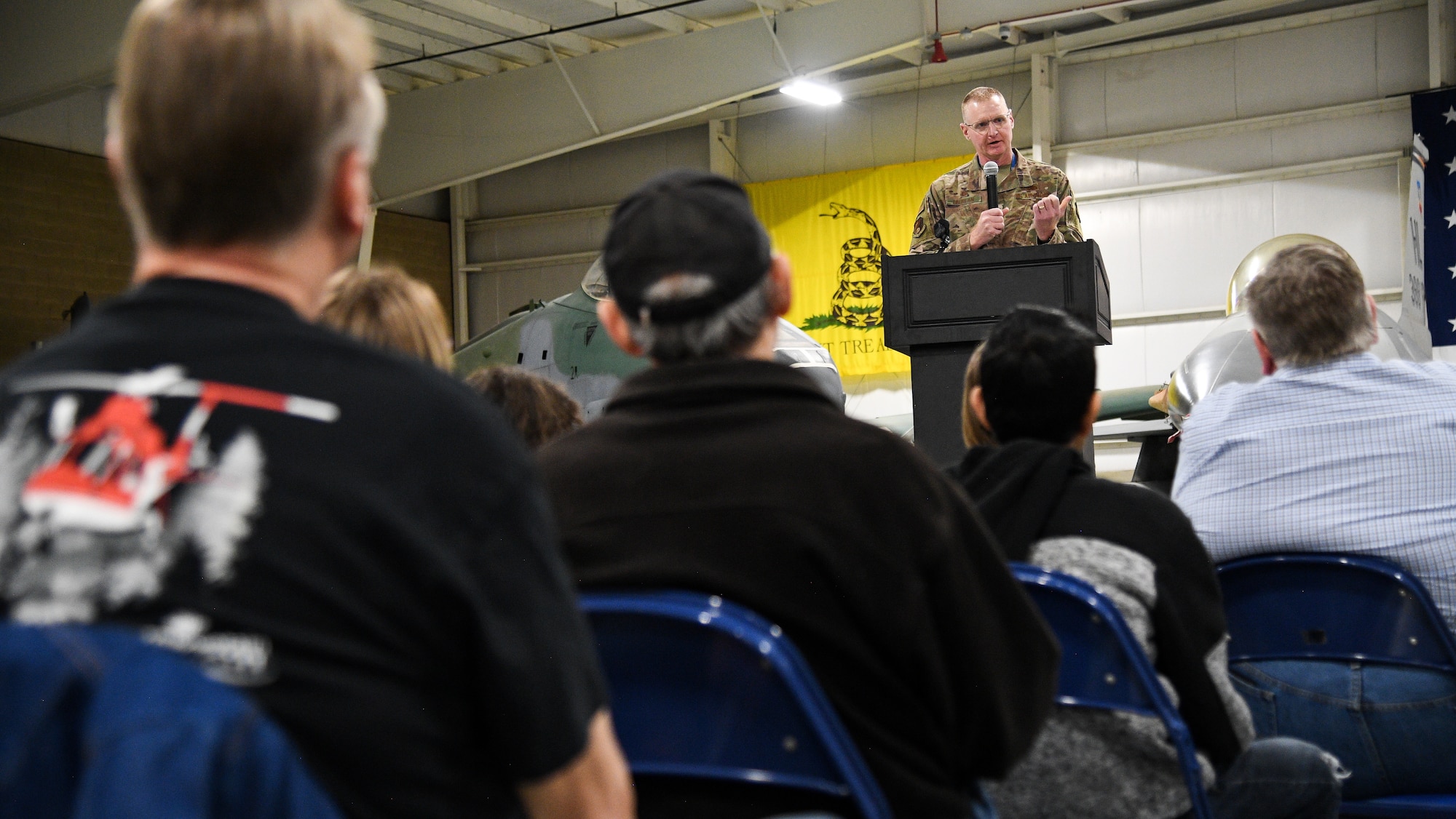 Col. Jon Eberlan stands behind a podium addressing members of an audience who appear in the foreground.