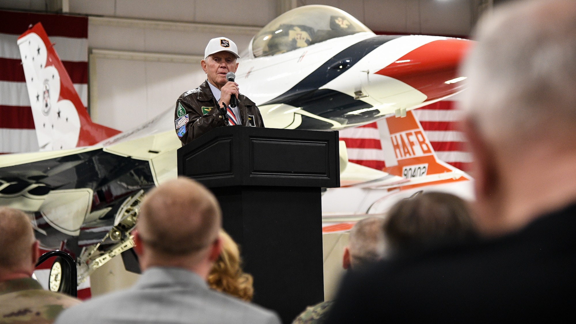 Retired Air Force Lt. Col. Jay Hess stands behind a podium in front of a static display of a U.S. Air Force Thunderbirds-painted F-16 inside the Hill Aerospace Museum. Audiences members appear in the foreground.