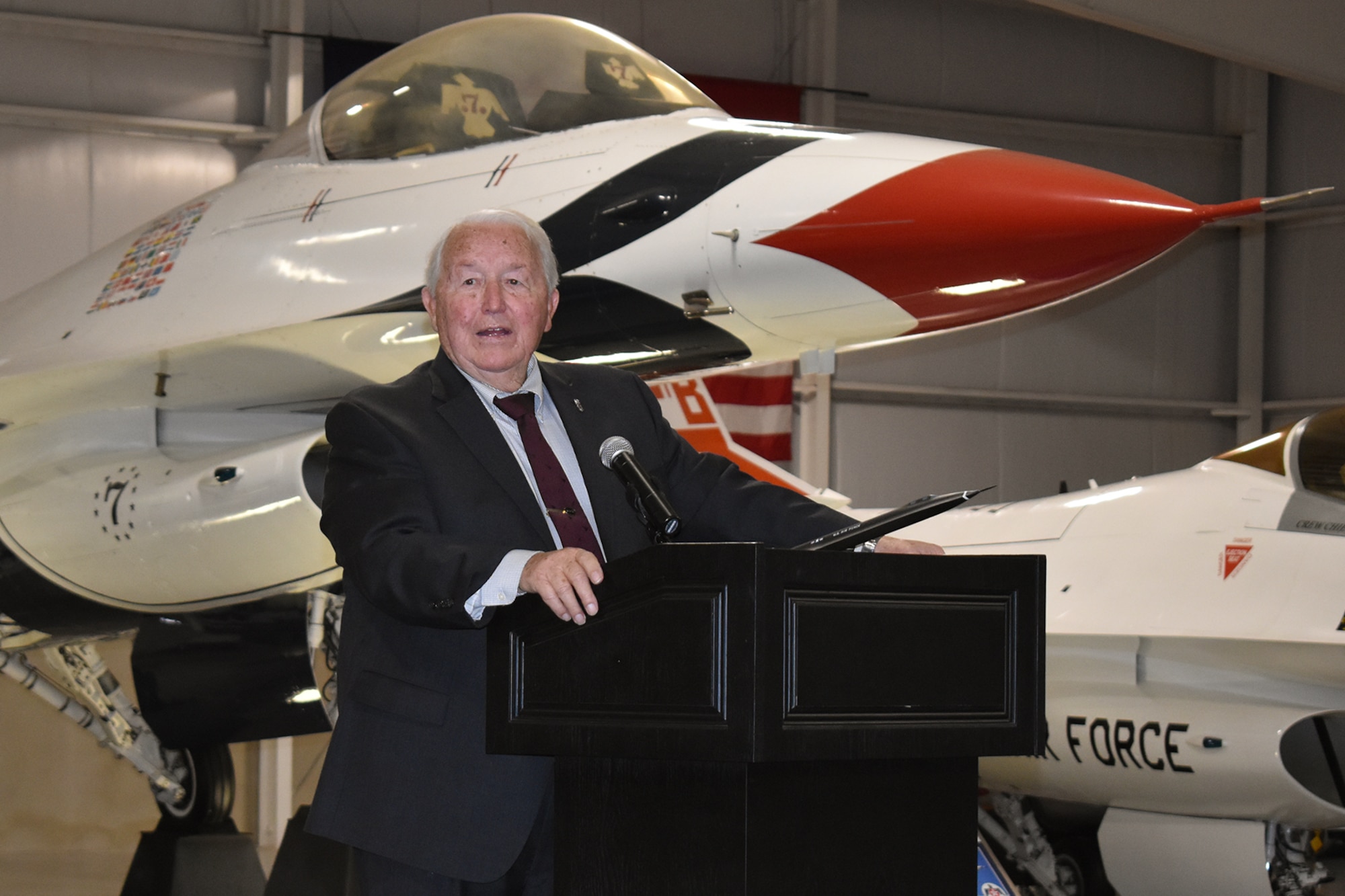 Retired Air Force Col. James Sullivan stands behind a podium located in front of a static display of a U.S. Air Force Thunderbirds-painted F-16 inside the Hill Aerospace Museum.
