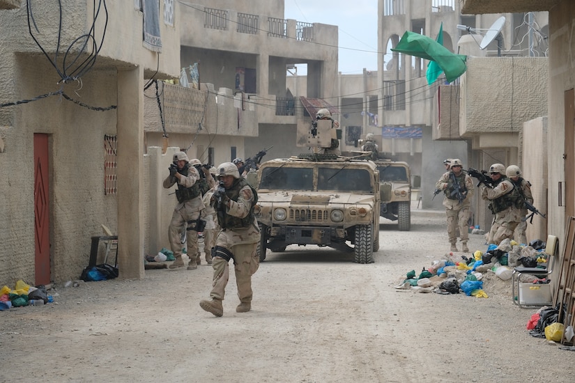 Several men dressed as soldiers walk with guns pointed on a set that looks like a street in Iraq. Two armed vehicles travel with them.