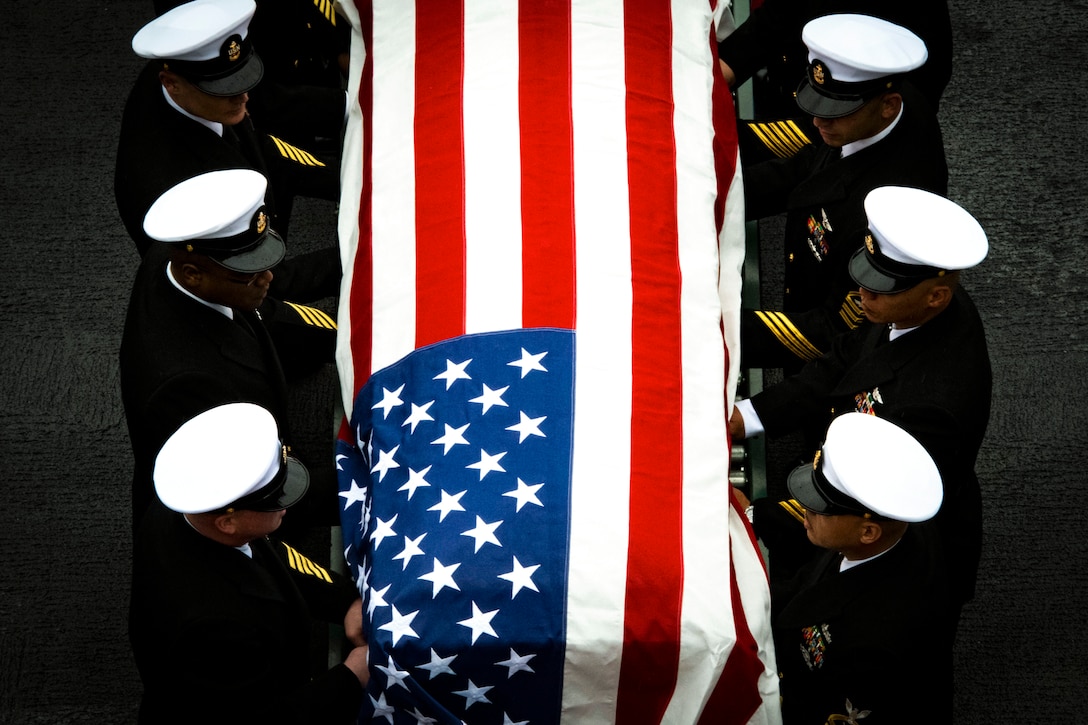 A group of sailors stand in line around a casket covered by the American flag.