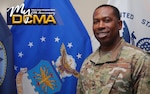 A smiling male in a military uniform stands beside a flag.