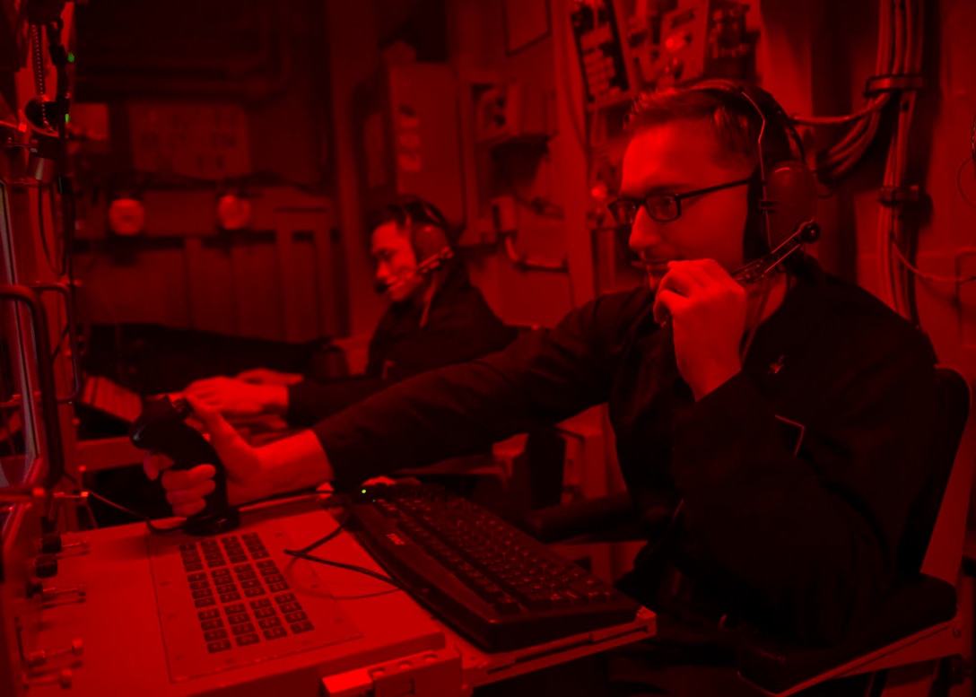Two sailors sit in a red control room on a ship.