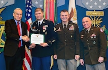 One male in suit shaking hands with male Soldier in blue dress uniform holding a certificate and two male Soldiers in green service uniform against a blue backdrop with flags and medal of honors painted on the wall.