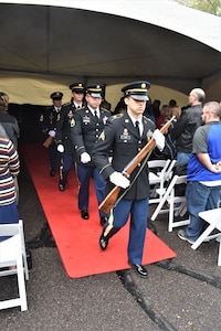 Soldiers in dress blue uniform with service cap and white gloves march down a red carpet in the middle of a crowd two hold rifles.