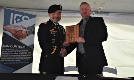 Male Soldier in blue dress uniform and beret shakes hands with male in suit while holding a wood plaque. An IES banner is standing to the left of the duo.