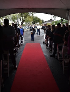People standing in chairs on both sides of a red carpet with male Soldiers in blue dress uniform with ceremony rifles standing at the end of the carpet.