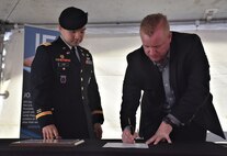 Male Soldier in blue dress uniform with beret stands next to and watches civilian man in dark suit signing a piece of paper with a pen.