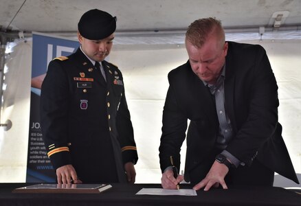 Male Soldier in blue dress uniform with beret stands next to and watches civilian man in dark suit signing a piece of paper with a pen.